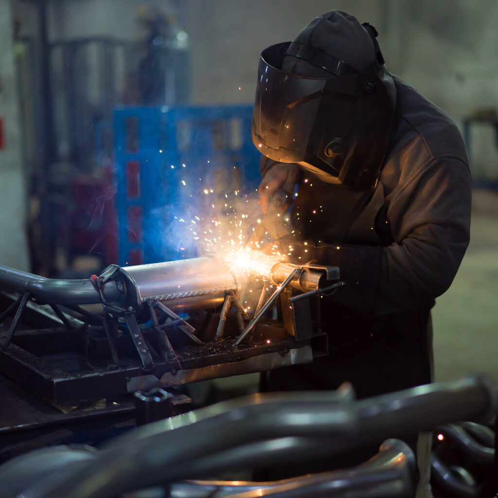 dramatic view of a worker wearing protective gear, welding together pieces of an exhaust pipe, with several other pipes around, on the work table, and metal blue frames in the background, in an industrial setting
