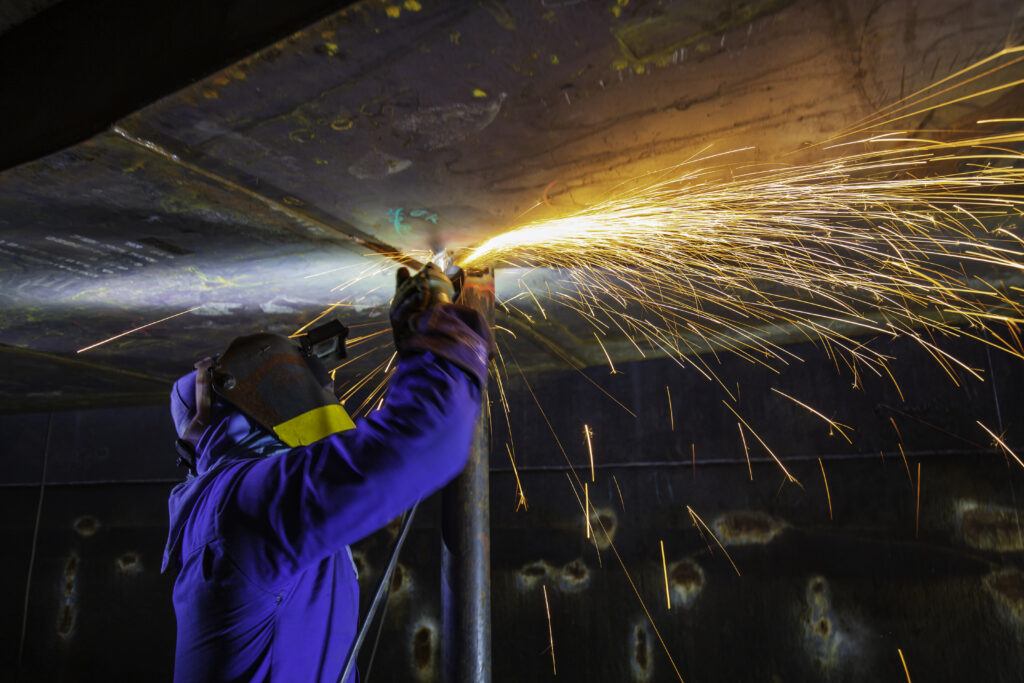 Male worker grinding on steel plate with flash of sparks close up wear protective gloves oil inside confined spaces.